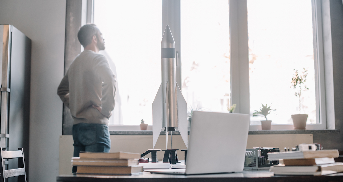 Pensive engineer looking at window, rocket model on tabletop at home